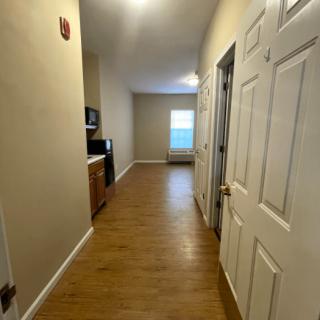 A long apartment hallway with wooden flooring, leading to a kitchen area and a window.