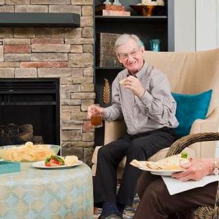 An elderly man enjoying snacks and a drink in a cozy living room by the fireplace.