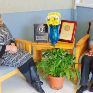 Two individuals seated indoors beside a table displaying awards, a plant, and a vase with flowers.