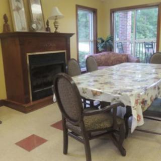 A cozy dining area featuring a table with a floral tablecloth, surrounded by chairs and near a fireplace with large windows for natural light.