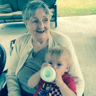 An elderly woman holds a young child who is drinking from a bottle outdoors.