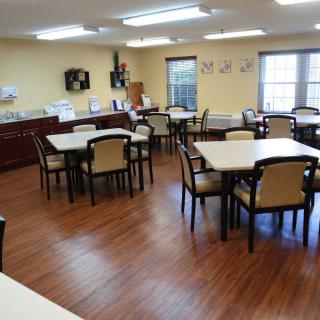 An empty dining room with neatly arranged tables and chairs, featuring a wooden floor and kitchen area.
