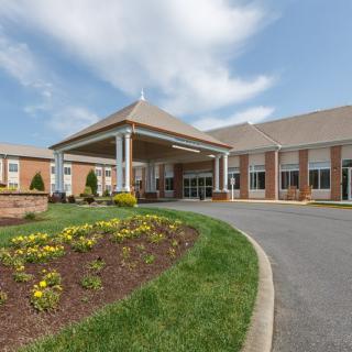 The image shows the entrance of a large building with a landscaped driveway and a partly cloudy sky.