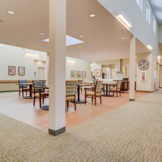 A spacious and well-lit dining area with tables and chairs, featuring pillars and a carpeted floor.