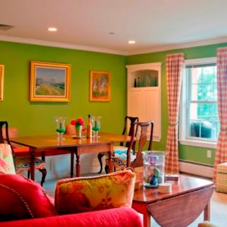 A cozy dining room with vibrant green walls, adorned with artwork and featuring a wooden dining table surrounded by chairs. The room is well-lit by natural light through a window with checkered curtains.