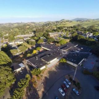 Aerial view of a complex of buildings surrounded by trees and hills with a parking lot in the foreground.