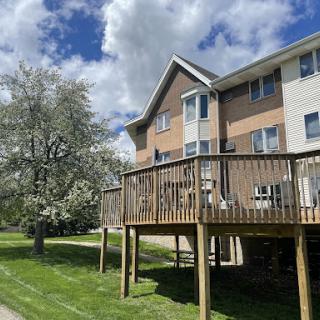 A residential building with a wooden deck, surrounded by green grass and a flowering tree under a partly cloudy sky.