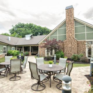 A cozy outdoor patio area with tables and chairs, set against a building with a stone facade and surrounded by lush landscaping.