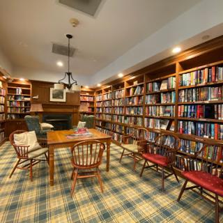 A cozy library featuring wooden bookshelves filled with books, a central table, and comfortable seating illuminated by warm lighting.