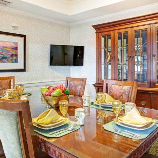 A well-appointed dining room featuring a polished wooden table set with colorful place settings, surrounded by elegant chairs and accented with a decorative china cabinet, wall-mounted TV, and artwork.