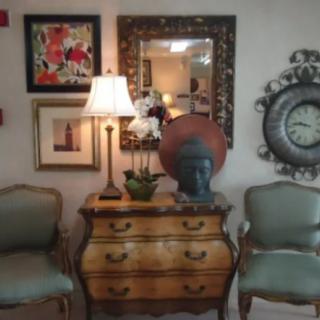A vintage-style interior with an antique dresser, decorative mirror, framed art pieces, a table lamp, and two elegant chairs.