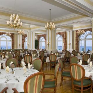 An elegant dining room with classic decor, featuring chandeliers and large windows offering a sea view.