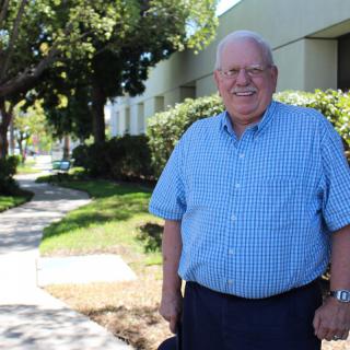 An older man smiling while standing on a sunny sidewalk lined with trees.