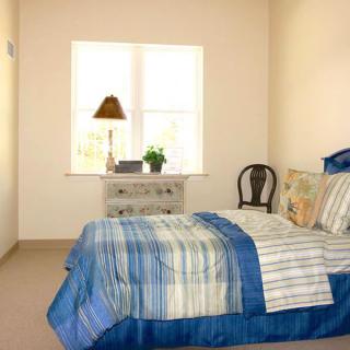 A cozy bedroom with a neatly made bed, a nightstand with a lamp and plant, and a window letting in natural light.