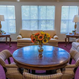 A cozy living room with a round wooden table, surrounded by chairs, two sofas, and large windows letting in natural light.