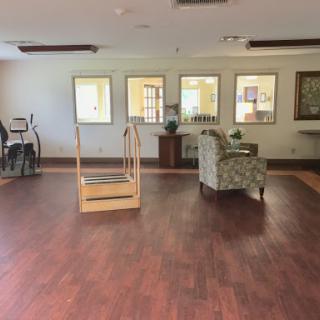 A spacious and empty lounge area featuring a wooden floor, a patterned chair, and rehabilitation equipment in the corner.