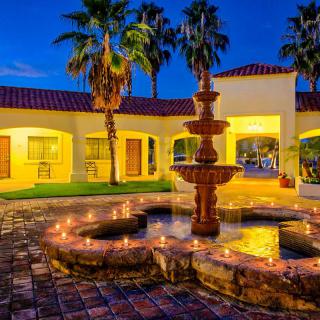 A beautifully lit courtyard with a central fountain illuminated at night, surrounded by palm trees and a building with warm lighting.