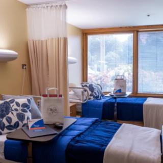 A well-lit hospital room featuring two neatly made beds with blue bedding and floral pillows.