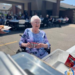 An elderly woman enjoys a sunny day at a community gathering, smiling as she receives food.