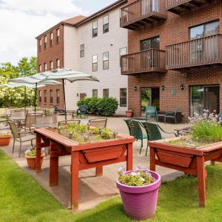 An outdoor garden area with raised planters, tables, and chairs under umbrellas beside a multi-story building.