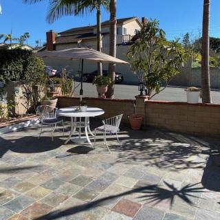 A sunny outdoor patio area with a table, chairs, and an umbrella surrounded by potted plants and palm trees.