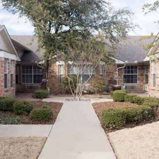 A brick residential house with landscaped bushes and trees, featuring a concrete sidewalk leading to the entrance.