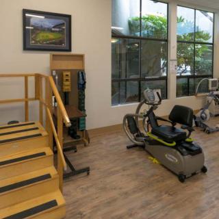A well-lit gym with exercise equipment including a recumbent bike and stairs for rehabilitation exercises.