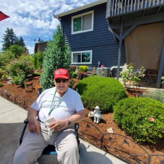 A man in a red cap sits in a wheelchair outside a house with a garden on a sunny day.