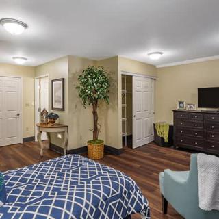 A cozy bedroom featuring a bed with a checkered blanket, a dresser with a TV on top, and decorative elements like a potted plant and armchair.
