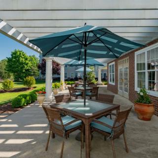 A sunny patio with outdoor seating and umbrellas under a pergola, surrounded by a garden.
