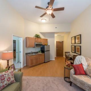 A cozy living room with a kitchenette and comfortable seating area, featuring warm decor and a ceiling fan.