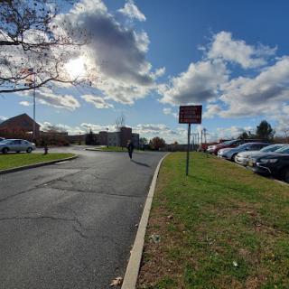 A lone figure walks along a winding road beside a parking lot under a partly cloudy sky.