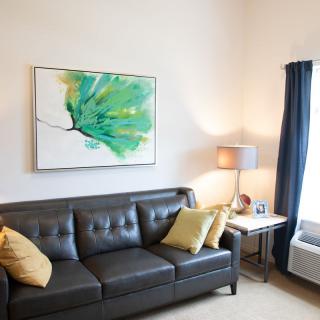 A modern living room with a dark leather sofa, colorful artwork, and a cozy lamp by the window.