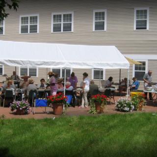 A group of people gathered under a white tent outside a house with a grassy lawn and flower arrangements.