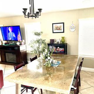 A well-lit dining room with a large table, chairs, and a TV on in the background.