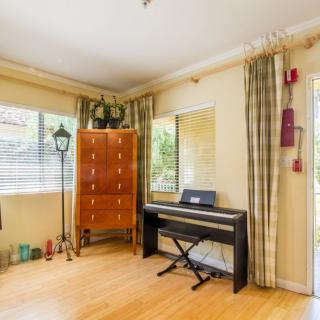 A cozy living room featuring a digital piano and a wooden chest of drawers against a bright, windowed wall with light curtains.