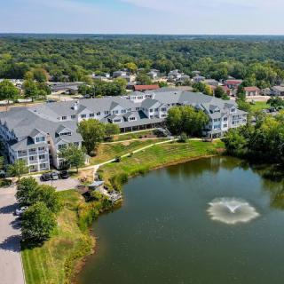 Aerial view of a residential building near a pond with a fountain, surrounded by trees in a suburban area.