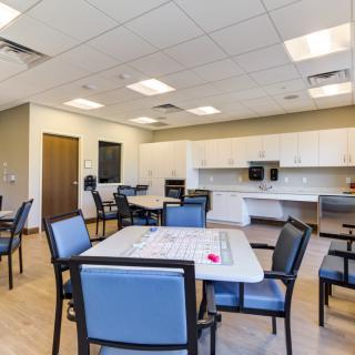 A modern indoor room featuring tables and chairs, with a kitchenette in the background.