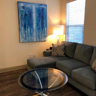 A stylish living room with a gray sectional sofa, glass coffee table, and blue abstract painting on the wall.