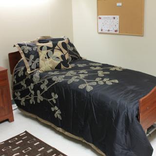 A cozy bedroom featuring a bed with dark floral bedding, complemented by a nightstand and a corkboard on the wall.