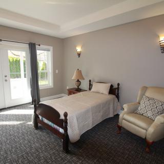 A cozy bedroom with a single bed and armchair, featuring a carpeted floor and natural light from a window and door.