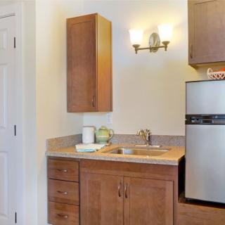 A small kitchen area featuring wooden cabinets, a sink, a countertop, and a compact fridge under warm lighting.