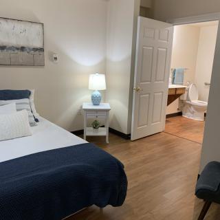 A cozy bedroom with a neatly made bed, nightstand with a lamp, and an open doorway leading to a bathroom.
