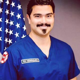 A man in blue medical scrubs stands in front of an American flag.