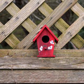 A small red birdhouse with white flower decorations sits against a wooden lattice fence.