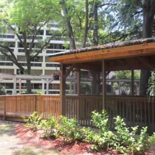 A wooden gazebo surrounded by lush greenery, with a modern building in the background.