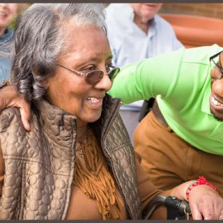 An elderly woman wearing sunglasses is smiling, sharing a warm moment with a friend outdoors.