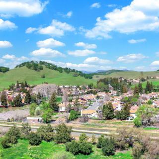 A scenic view of a lush green landscape with rolling hills and a quaint village under a bright blue sky dotted with clouds.