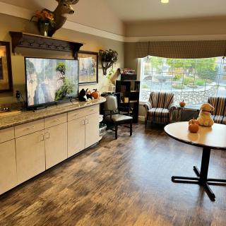 A cozy living room with a large television, seating area, and a table near a bright window.