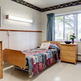 A hospital room with a single bed, decorated with a colorful quilt, alongside wooden furniture and a large window.